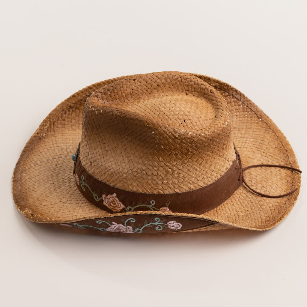 Tea colored straw cowboy-shaped hat with decorative floral pattern band and underbrim on a light beige background