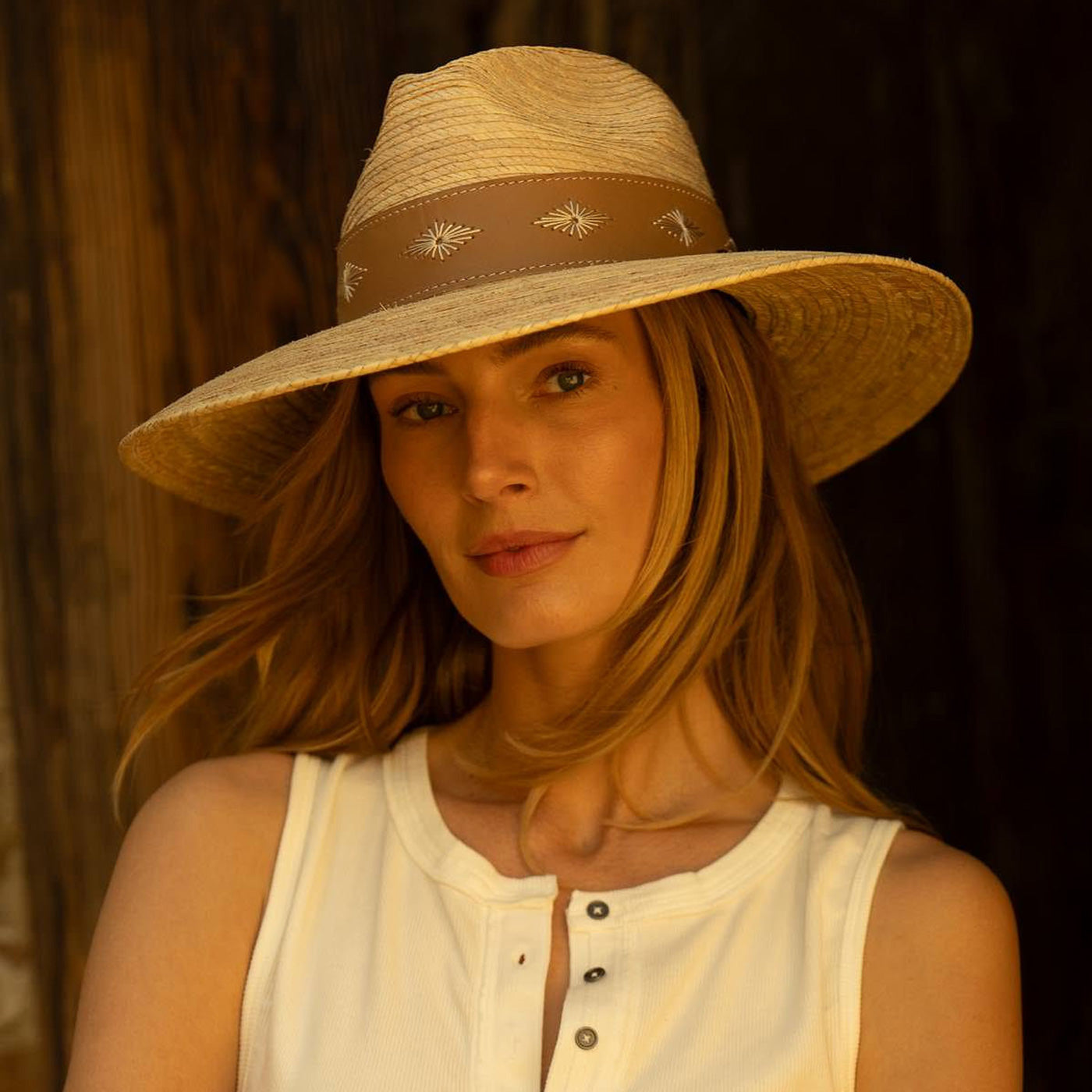Woman wearing a wide-brimmed straw hat with a decorative dark brown band standing against a dark wooden background