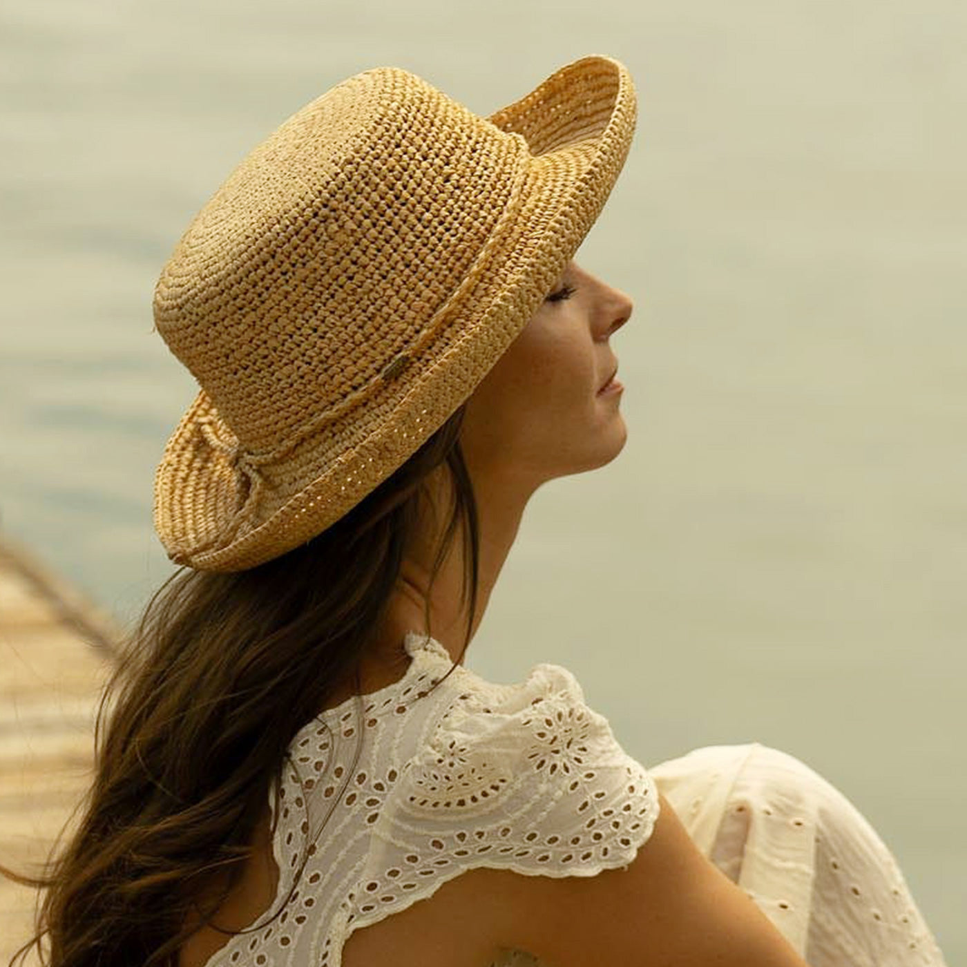 Woman facing upwards wearing a straw hat and white lacey dress sitting on a wooden dock with a blurred natural background