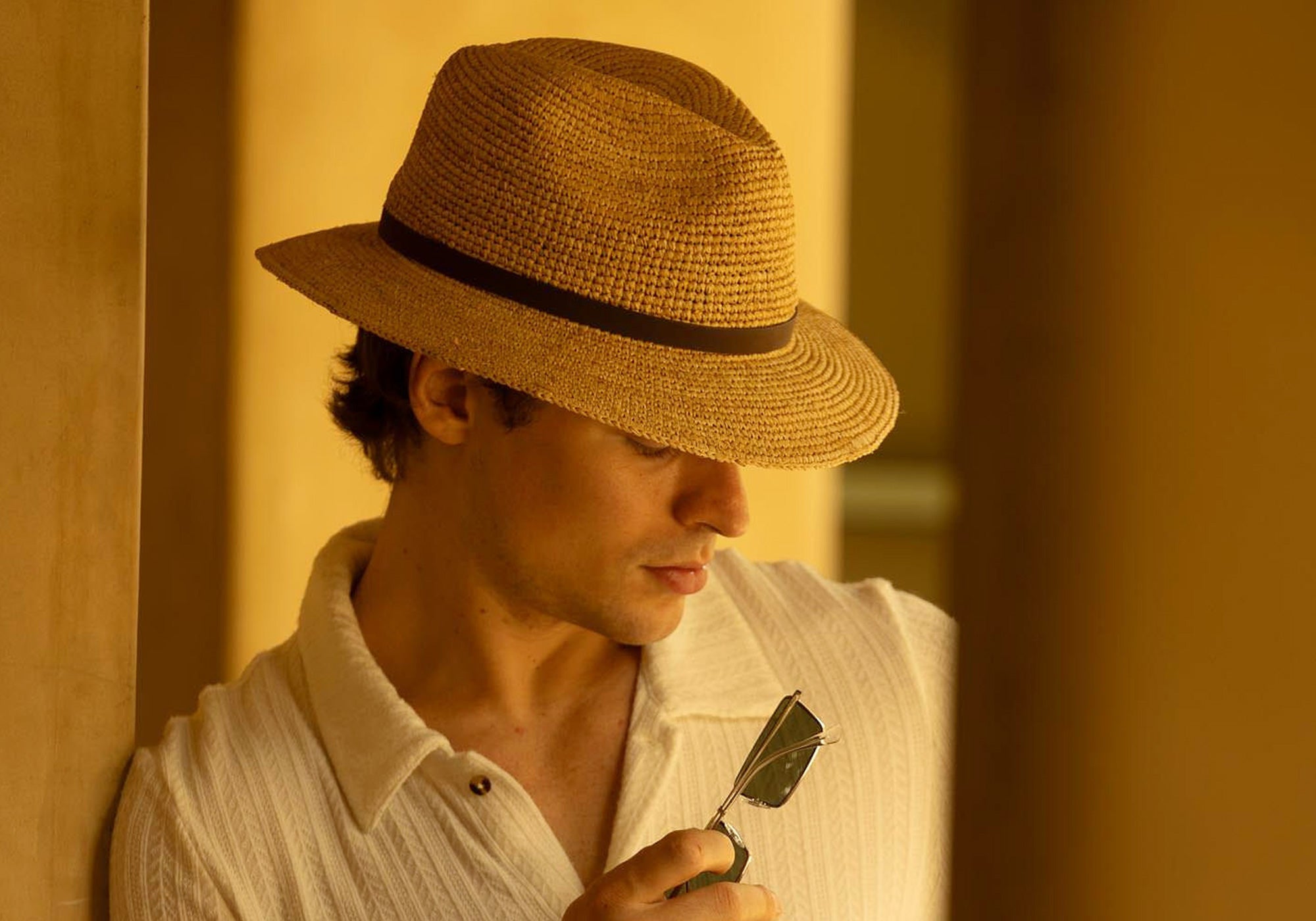 Man wearing a golden colored straw hat and white shirt holding a pair of sunglasses in a warm-toned setting, looking downward towards the right
