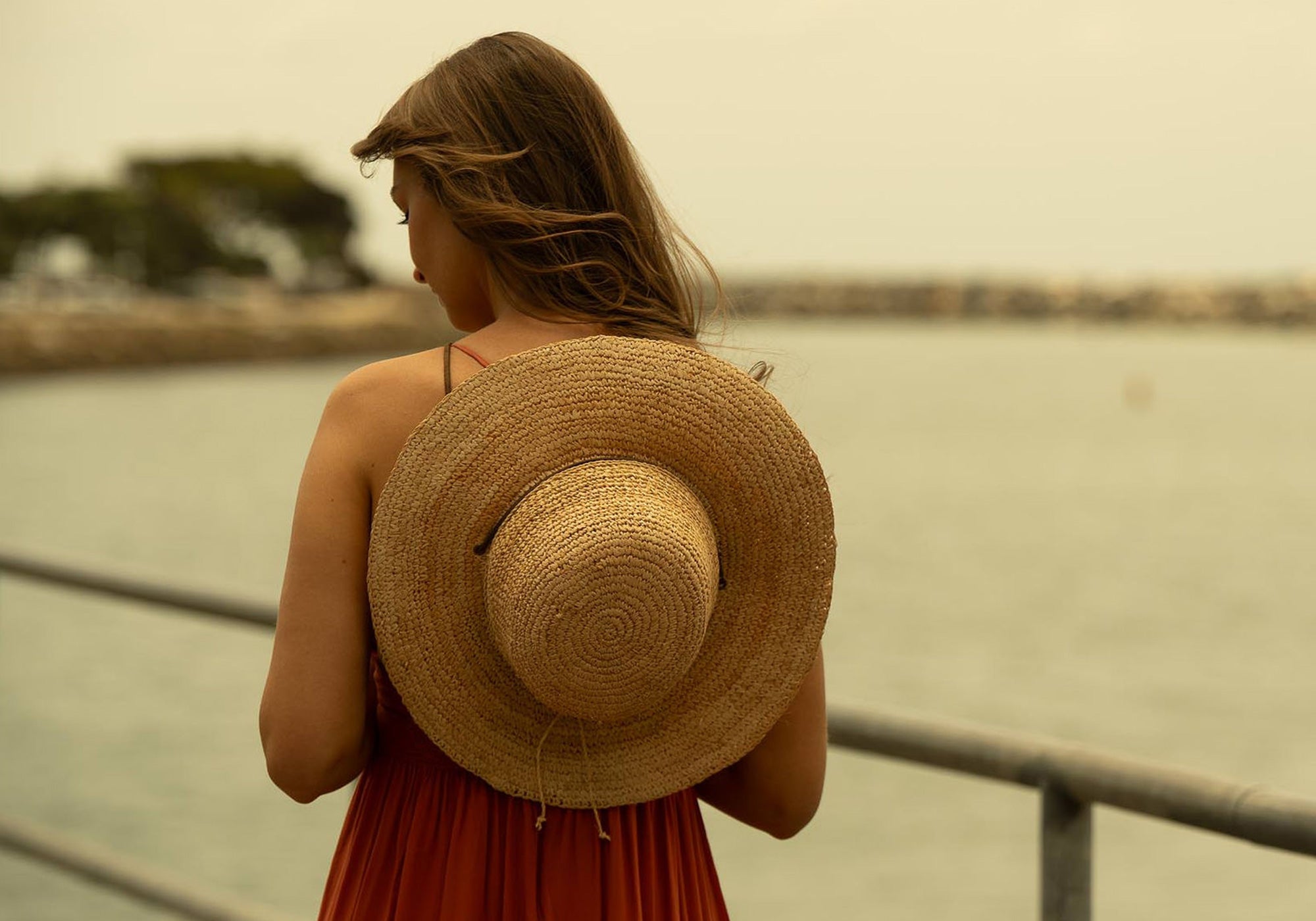 Woman in a red dress with straw hat resting on her back by its drawstrings standing facing away next to body of water with a blurred background
