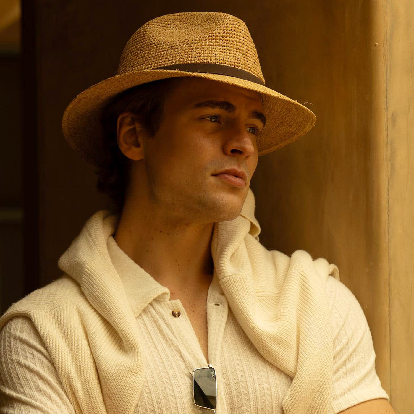 Man wearing a beige straw hat and light-colored shirt with sunglasses hanging off the front, standing against a warm-toned background