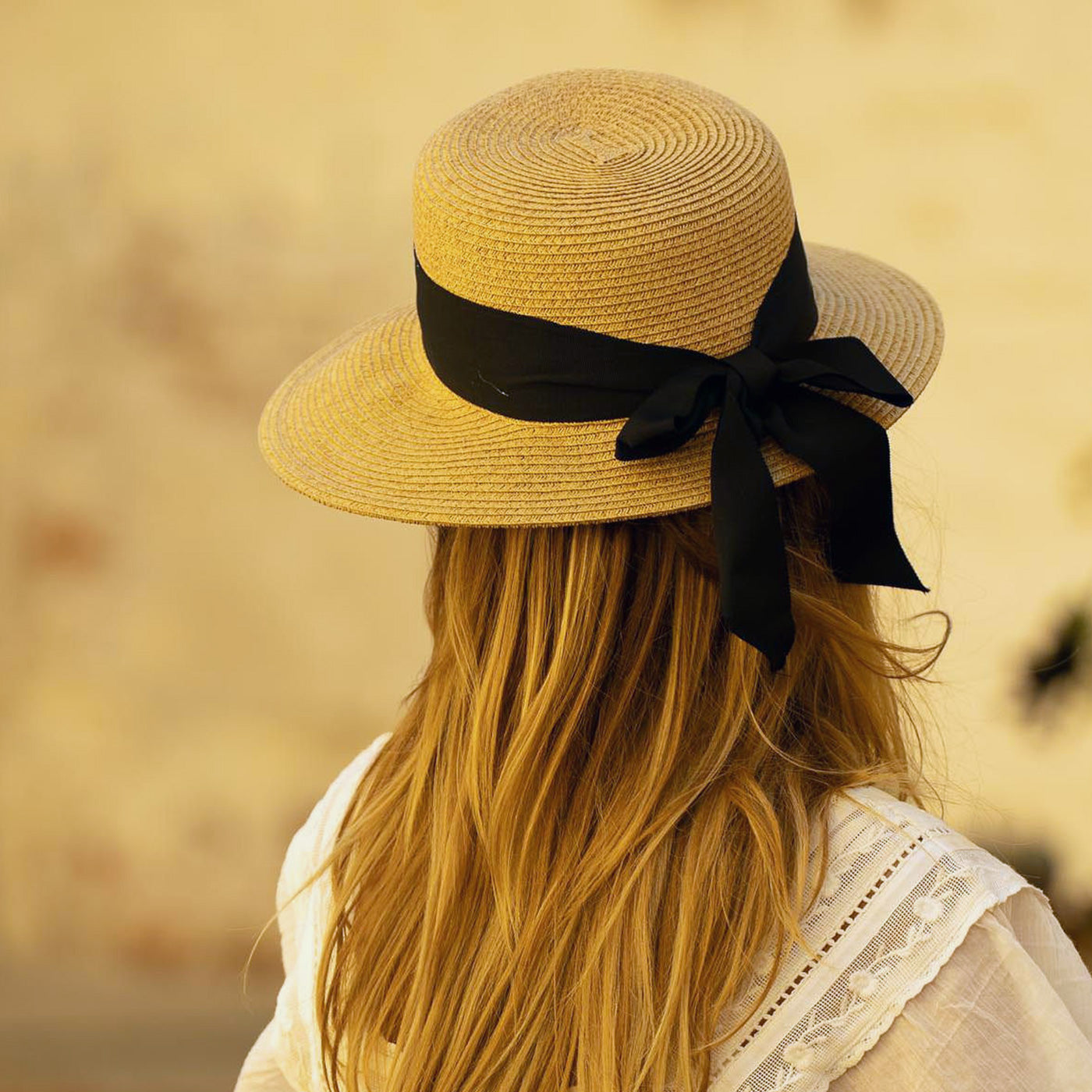 Woman wearing a straw hat with a black thick ribbon and a white lacy dress facing away against a blurred background